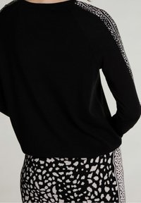 Schwarzer Pullover mit einer strukturierten Oberfläche, mit gemusterten Streifen entlang der Schultern. Kombiniert mit schwarz-weiß gepunktetem Stoff.