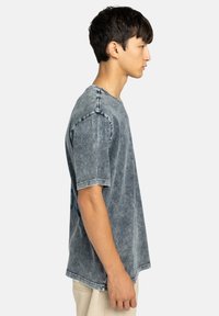 Grijze denim t-shirt met korte mouwen, relaxed fit, zichtbare stiksel details en een verwoeste textuur. Zijprofielweergave.