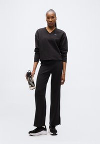 Γυναίκα με μαύρο φούτερ και παντελόνι Puma, κρατώντας ένα διαφανές μπουκάλι νερού, στέκεται σε λευκό φόντο.