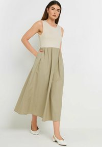 Beige ärmelloses Kleid mit einem fließenden olivgrünen Rock, der über Seitentaschen und einen einfachen runden Ausschnitt verfügt. Insgesamt glatte Textur.