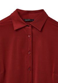 Camicia rossa con colletto, dal tessuto liscio, con chiusura frontale a bottoni e bottoni coordinati. Etichetta visibile con il marchio "CALLIOPE".