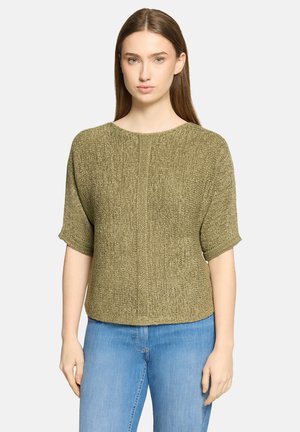 Vrouw met stijl bruin haar, draagend een olijfgroene gebreide trui met korte mouwen en lichtblauwe jeans, staand tegen een eenvoudige achtergrond.