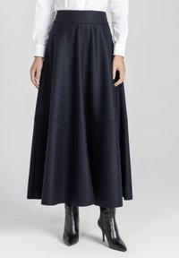 Kobieta w długiej czarnej spódnicy w kształcie litery A z bocznymi kieszeniami, białej koszuli z długim rękawem oraz czarnych botkach na wysokim obcasie z wysmuklonym noskiem.