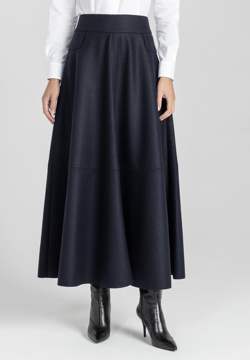 Kobieta w długiej czarnej spódnicy w kształcie litery A z bocznymi kieszeniami, białej koszuli z długim rękawem oraz czarnych botkach na wysokim obcasie z wysmuklonym noskiem.