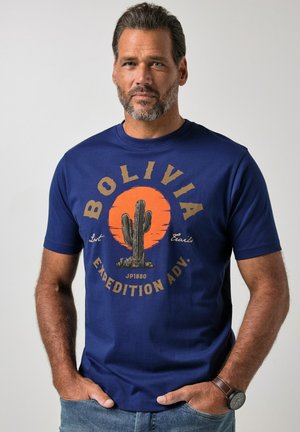 Uomo con maglietta blu con scritta marrone "Bolivia Expedition Adv." e grafica di un cactus davanti, in piedi con le mani nelle tasche.