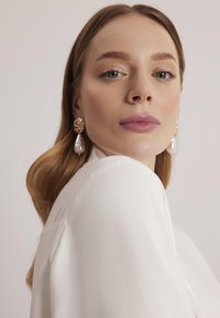 Złote kolczyki z teksturowanymi okrągłymi końcówkami i białymi perłami w kształcie kropli; modelka ma na sobie gładką białą odzież. Miękkie, neutralne tło.