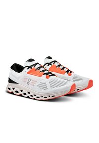 Hvide atletiske sko med sorte accenter og orange detaljer. Mesh-overdel med et struktureret mønster og en polstret sål med udskæringer.