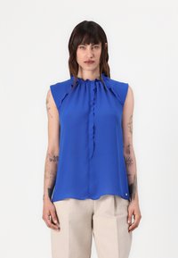 Blauwe mouwloze blouse met een gerimpelde nekdetail en een gladde textuur; de zoom valt net onder de taille. Gecombineerd met lichtbeige broeken.