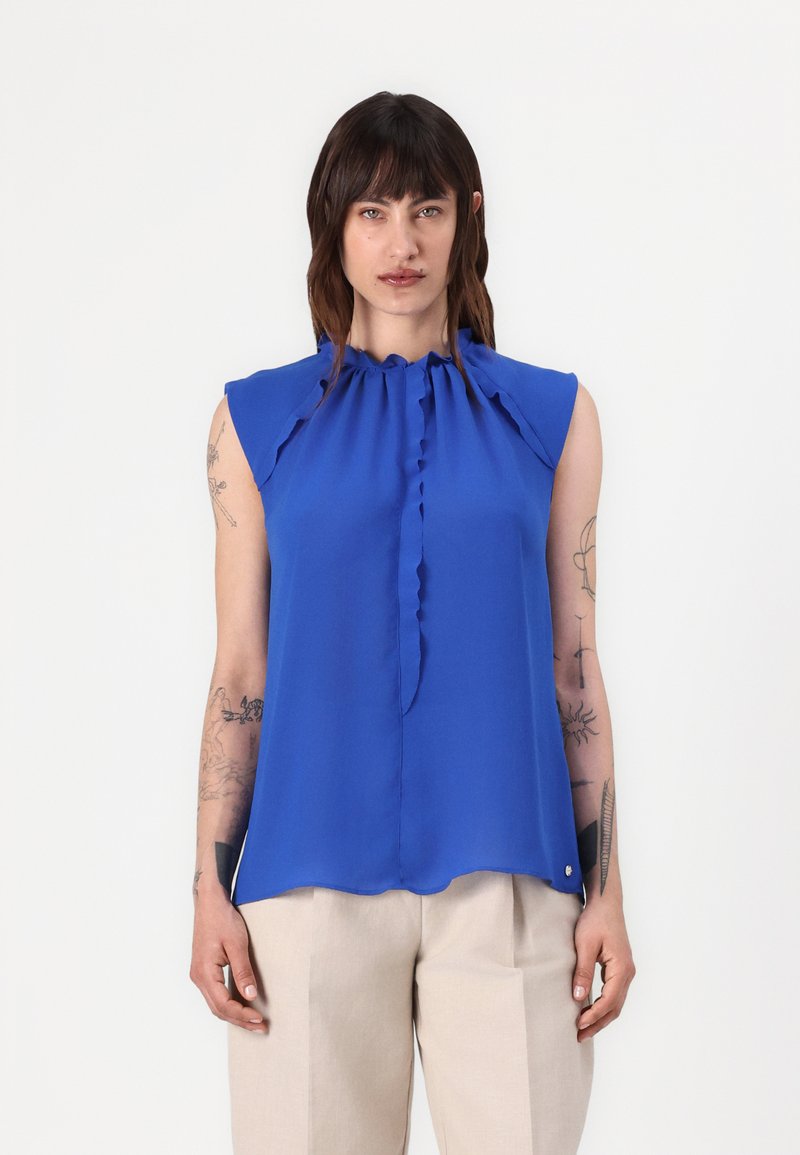 Blauwe mouwloze blouse met een gerimpelde nekdetail en een gladde textuur; de zoom valt net onder de taille. Gecombineerd met lichtbeige broeken.