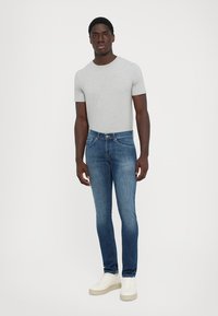 Homme portant un t-shirt à manches courtes gris clair, un jean bleu slim et des baskets blanches, se tenant contre un fond blanc uni.
