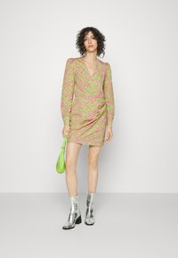 Vestido curto e ajustado em estampa de leopardo vibrante verde e rosa, com mangas longas, decote em V profundo e drapeado lateral. Botas prateadas até o tornozelo e uma pequena bolsa verde completam o visual.