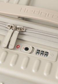 Gros plan sur la fermeture éclair beige d'une valise avec doubles tirettes, verrou TSA, molettes de combinaison réglées sur 000, et le nom de la marque « Hedgren » sur la poignée.