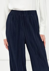 Femme portant un pantalon large plissé bleu marine et un chemisier blanc à manches longues, les mains reposant le long du corps.