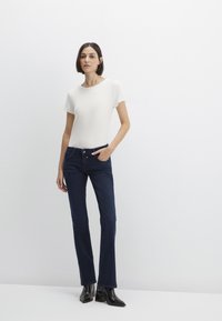 White ribbed short-sleeve top paired with dark blue flared jeans. Model stands with one hand in pocket, wearing black ankle boots.