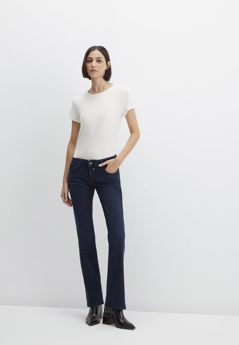 White ribbed short-sleeve top paired with dark blue flared jeans. Model stands with one hand in pocket, wearing black ankle boots.