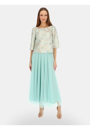 Kobieta w jasnoniebieskiej bluzce w kwiatowy wzór z rękawami do połowy łokcia oraz płynącej, plisowanej długiej spódnicy w tym samym kolorze.