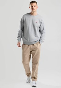 Sweatshirt gris à col rond, avec des poignets côtelés et un petit logo rouge, associé à un pantalon beige ample et des baskets blanches.