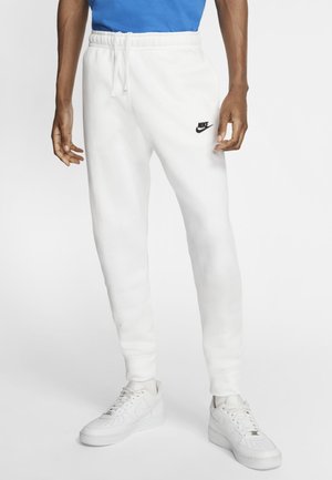 Mężczyzna w białych spodniach dresowych Nike z czarnym logo, białych butach sportowych i niebieskiej koszulce, stojący na tle jednolitego białego tła.