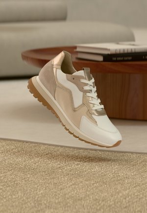 Beige, witte en metallic gouden sneaker met een gestructureerde zool die zweeft boven een geweven kleed in een modern woonkamerinterieur.