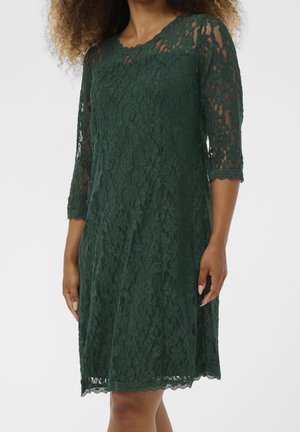 Mørkegrøn knælængdeblondekjole med trekvartærmer båret af en person med krøllet hår, stående mod en ensfarvet baggrund.