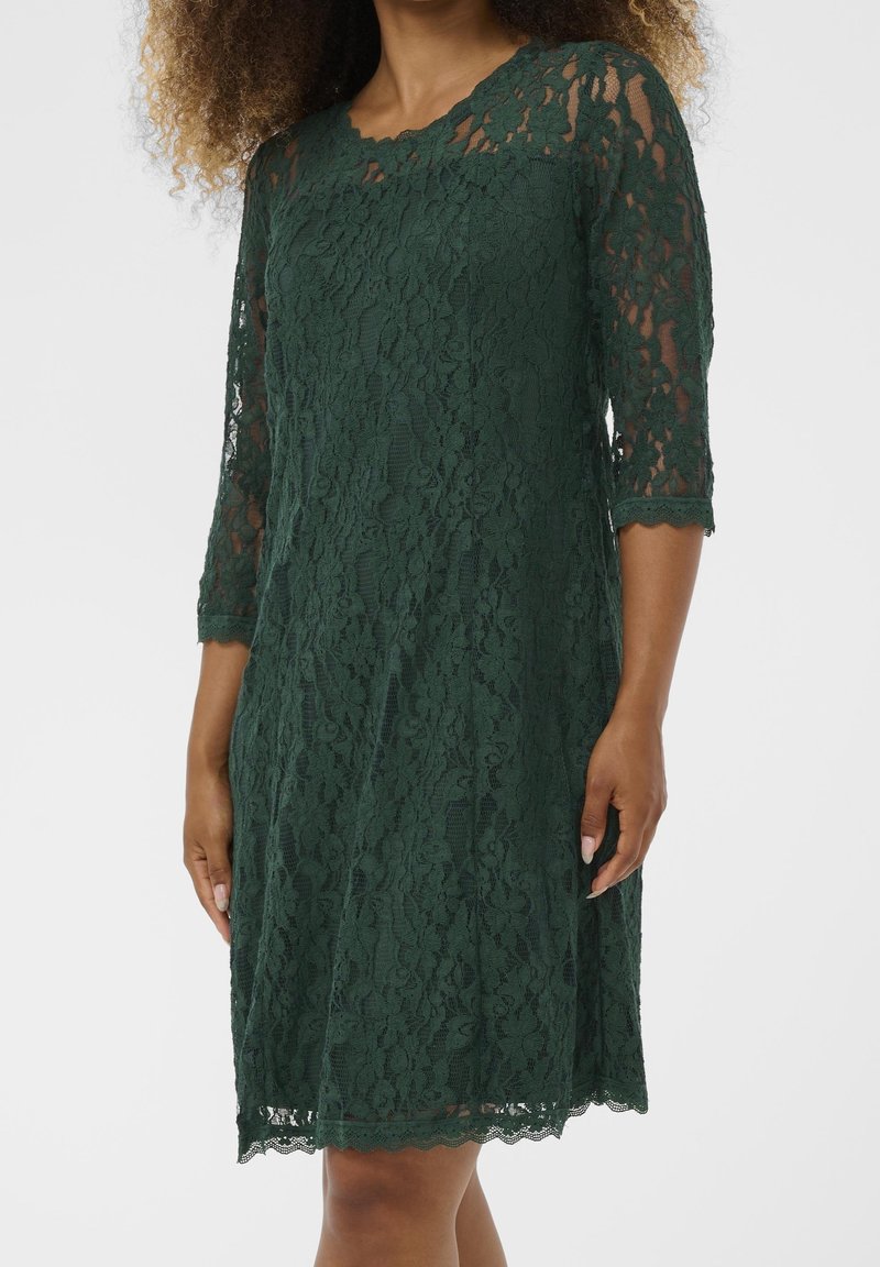 Robe en dentelle vert foncé longueur genou avec manches trois-quarts portée par une personne aux cheveux bouclés, debout devant un fond uni.