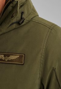 Maslinasto zeleni jakna s kapuljačom, teksturirani materijal, vidljivo šivanje i pravokutna smeđa oznaka s izvezenim motivom krila.