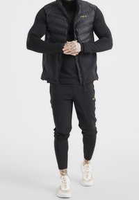 Černá vyp padded vesta přes černé dlouhé tričko, v kombinaci s černými tepláky a bílými teniskami. Minimalistický design s přiléhavým střihem.