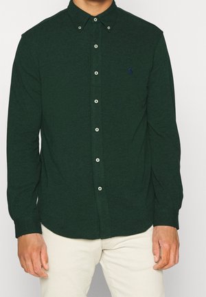 Mann trägt ein dunkelgrünes, geknöpftes Langarmhemd mit gesticktem Logo auf der Brust und helle beigen Hosen vor einem einfarbigen Hintergrund.