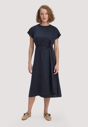 Frau mit lockigem Haar, die ein dunkelblaues midikleid mit kurzen Ärmeln und Taillengürtel trägt, kombiniert mit beigen, gewebten flachen Schuhen.