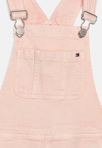 Overoles de mezclilla rosa claro con tirantes ajustables de metal, un bolsillo frontal con detalles de costura y una pequeña etiqueta de logo cerca de la costura del bolsillo.