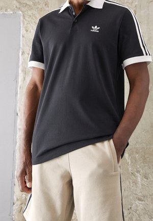 Czarna koszulka polo z białym kołnierzykiem i trzema paskami na rękawach, z małym logo Adidas na piersi; materiał bawełniany, luźny krój.