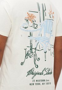 Flødefarvet bomulds-T-shirt med en blå og grøn linjetegning af et bord med blomster, kopper og en stol på bagsiden.