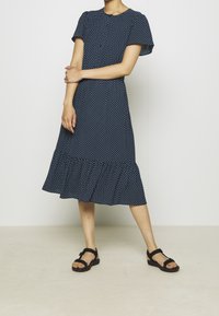 Žena stojící v námořnickomodré midi šaty s bílými puntíky a černými sandály na otevřenou špičku na jednoduchém bílém pozadí.