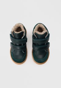 Dunkelgrüne Lederschuhe mit zwei Klettverschlüssen, weicher beiger Innenausstattung, kontrastierenden Wildlederakzenten und einer sandfarbenen Gummisohle.