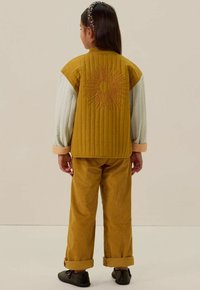 Gequiltede mosterdkleurige jas met een zonnestraalborduursel op de rug, gecombineerd met bijpassende corduroy broek en zwarte schoenen. Armbanden om de polsen.