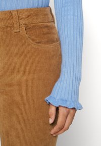 Ruka odpočívá na boku a má na sobě světle modrý žebrovaný svetr s volánkovými manžetami a hnědé kordové kalhoty s přední kapsou.