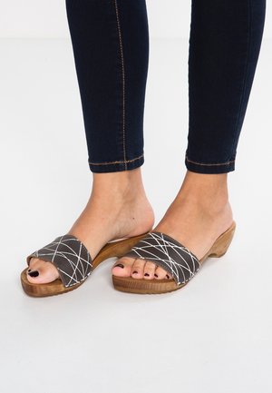 Jalat yang mengenakan sandal slip-on dengan tumit kayu dan tali bergaris hitam-putih, serta kuku jari kaki yang dicat gelap, berpakaian jeans ketat gelap.