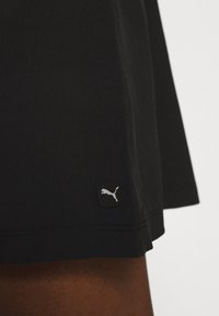 Jupe de sport noire en tissu doux, présentant un design droit et un petit patch logo Puma gris près de l'ourlet.