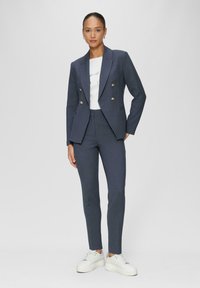 Blazer ajusté bleu marine avec des boutons dorés, associé à un pantalon slim et un t-shirt graphique blanc. Le modèle porte des baskets blanches chunky.