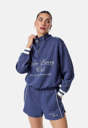 Kvinde iført navyblå sweatshirt med hvid tekst "Cardio Bunny Club" og matchende shorts, poserer med den ene hånd nær kraven.