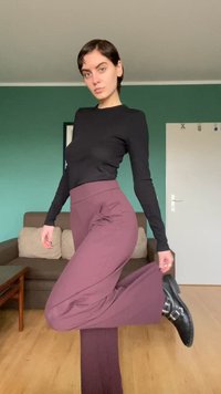 Zwart top met lange mouwen, bordeauxrode hoge taille broek met zijzakken, glanzende zwarte enkellaarzen met zilveren gespen. Achtergrond is een groene muur.