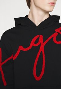 Schwarzer Hoodie mit einem auffälligen, strukturierten, roten gestickten Schriftzug. Weiches Baumwollmaterial mit einer klassischen Kapuzenform.