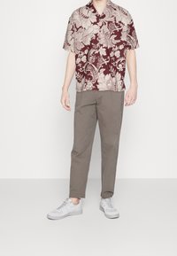 Camicia corta bordeaux con motivo floreale crema, abbinata a pantaloni tapered grigio chiaro e sneakers grigie, con un design semplice.