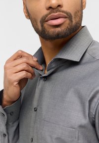 Graues Hemd mit Knopfleiste und strukturiertem Kragen sowie Manschetten. Die Knöpfe sind schwarz, und eine Brusttasche ist sichtbar. Der Stoff wirkt glatt mit leichtem Glanz.