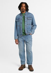 Άνδρας με dreadlocks που φοράει ανοιχτό μπλε τζιν μπουφάν και τζιν παντελόνι, πράσινο πουκάμισο και καφέ αρβυλάκια με κορδόνια, στέκεται με το ένα χέρι στην τσέπη.