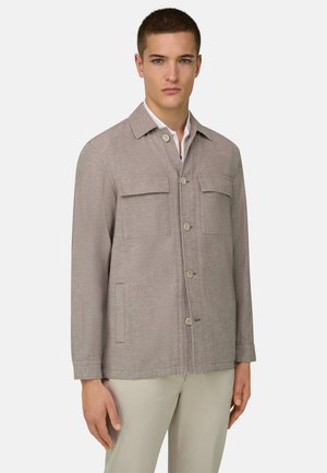 Joven vestido con una chaqueta beige abotonada con bolsillos con solapa en el pecho y bolsillos laterales con aberturas, combinada con pantalones de color claro.