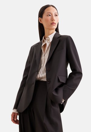 Femme aux longs cheveux foncés portant un blazer noir, un pantalon noir et une chemise blanche rayée boutonnée, debout avec une main dans la poche.