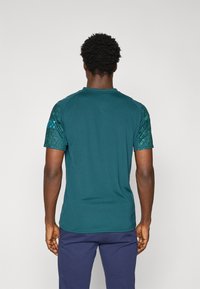 T-shirt à manches courtes en teal avec une texture lisse, présentant des accents à motifs sur les épaules. Encolure ronde, coupe décontractée et sans logos visibles.