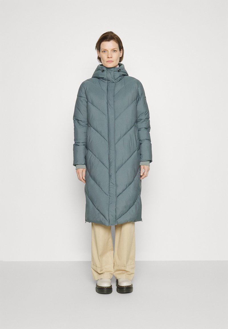 Mujer vestida con un abrigo acolchado largo gris con capucha, pantalones de pierna ancha beige y botas de plataforma blancas, de pie contra un fondo blanco simple.