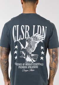 Muškarac s tetovažama nosi tamnosivu majicu s bijelom grafikom orla, zvijezdama i natpisima "CLSR LDN" i "Creators of Modern Essentials."